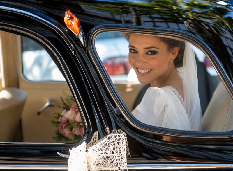 La novia sonriendo desde la ventanilla de un coche clasico el dia de su boda