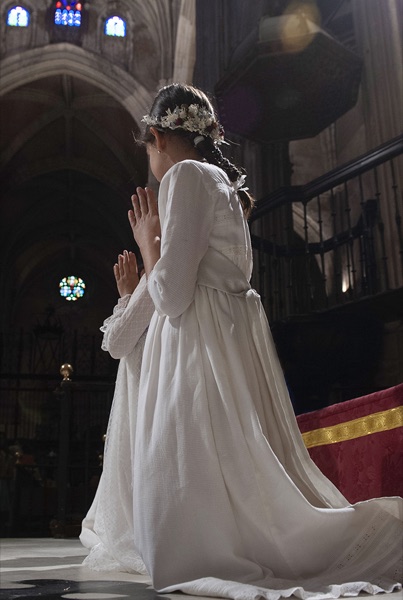 Nina de comunion arrodillada rezando de espaldas en la catedral, con corona de flores
